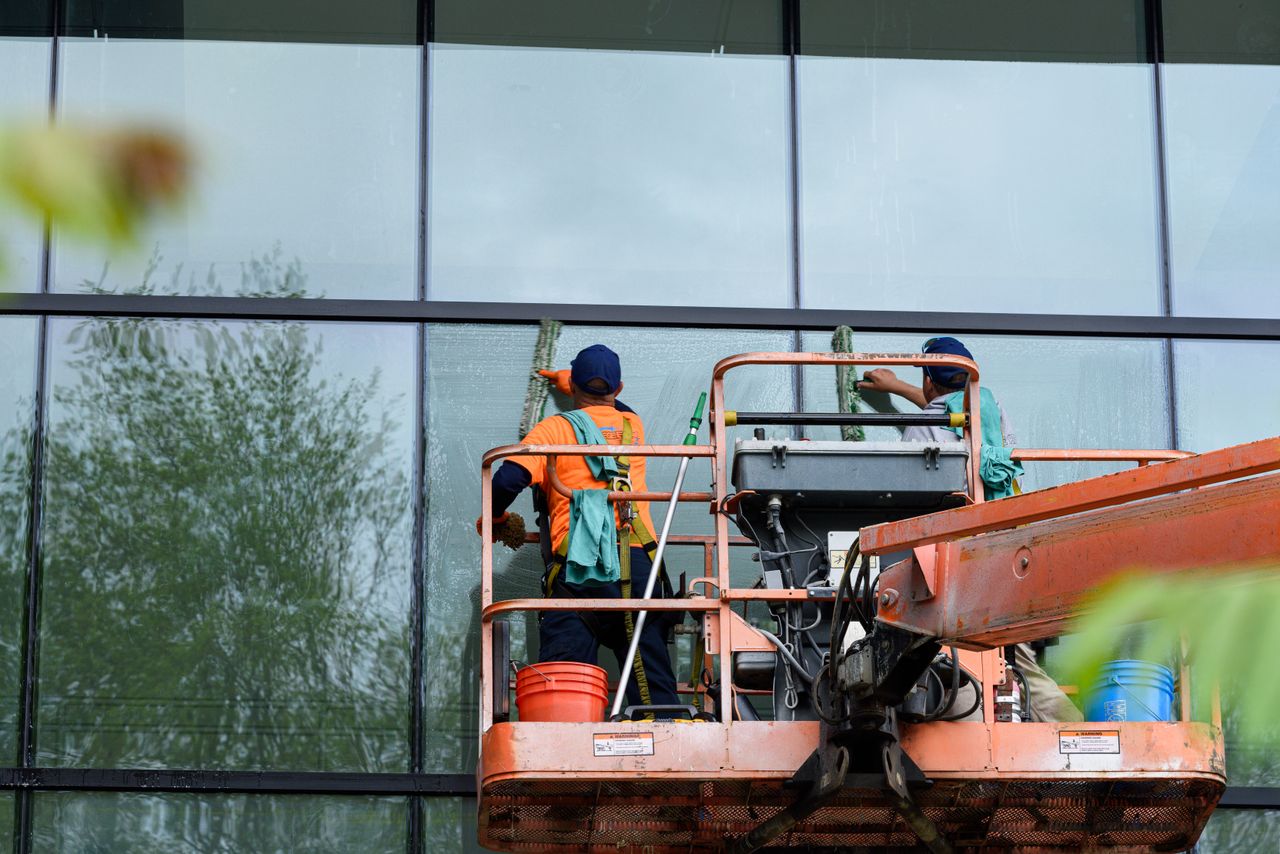 our-story Two workers washing windows on a glass building facade from a lift platform, improving the building’s appearance.